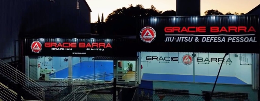 Fachada da Gracie Barra Santa Felicidade à noite, com letreiro vermelho iluminado e o tatame azul visível pelas vitrines em Curitiba